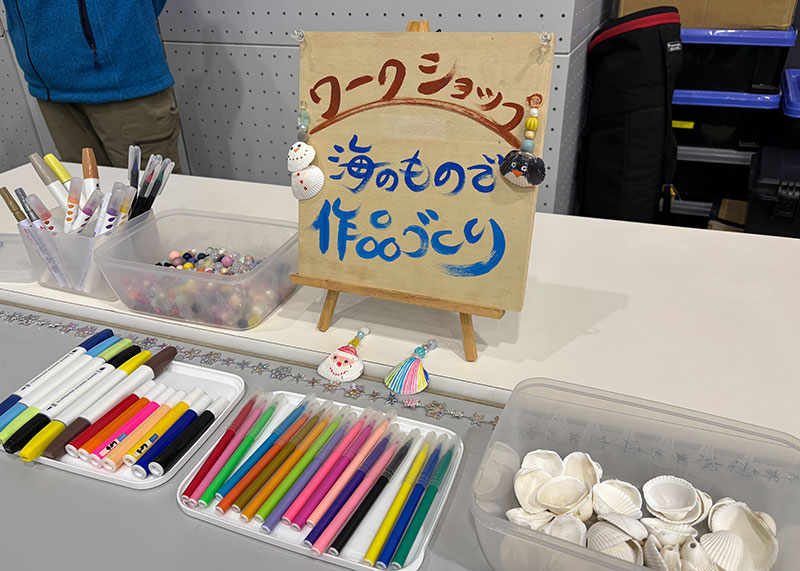 
										    「ワークショップ 海のもので作品づくり」と書かれたボードを中心に、
										    たくさんのカラーペンと貝殻が並んでいます
										  